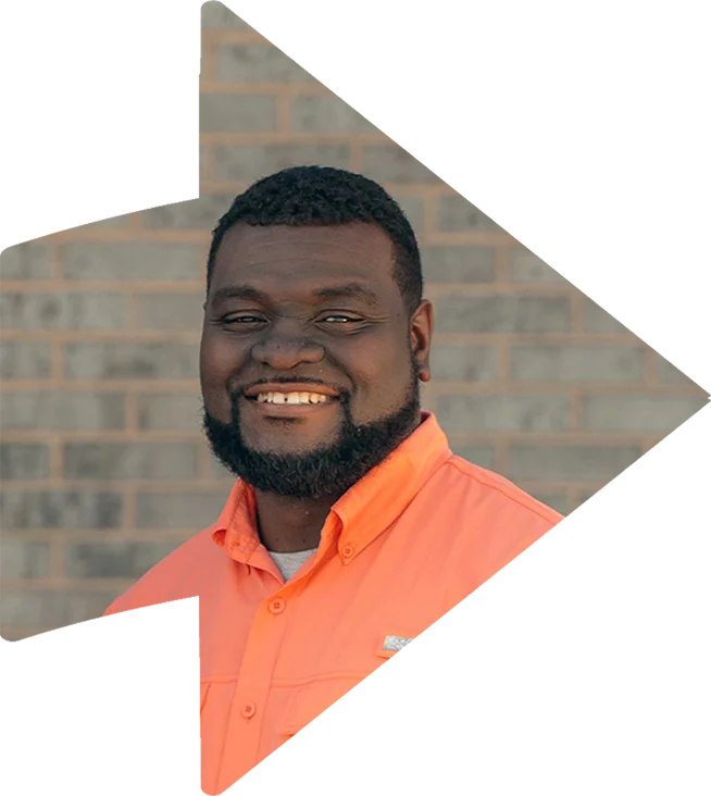 A man with a beard wearing an orange shirt smiles in front of a brick wall, framed by a large arrow graphic.