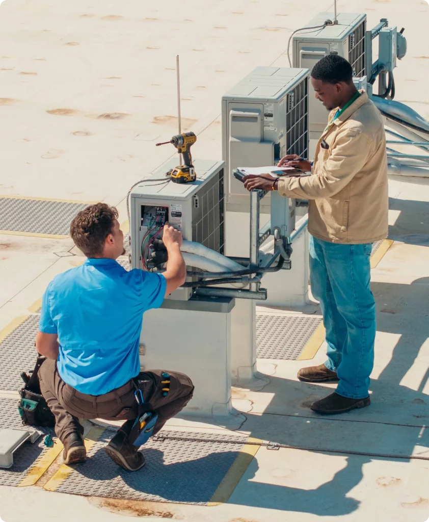 Two technicians work on rooftop HVAC units; one kneels and adjusts wiring while the other stands and references a tablet.