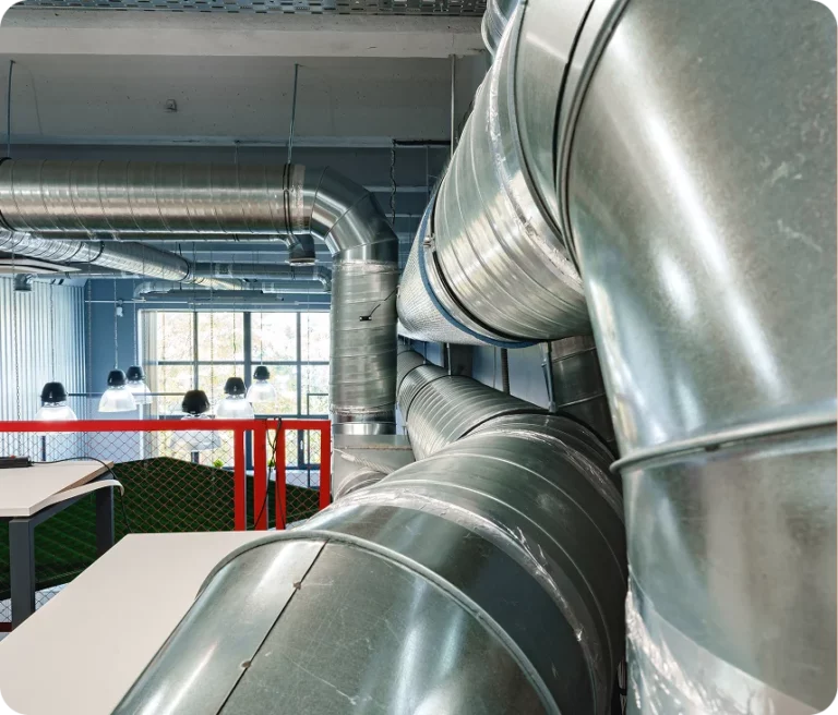 Large metal air ducts run through the interior of a modern industrial building with tables, chairs, and large windows visible in the background.