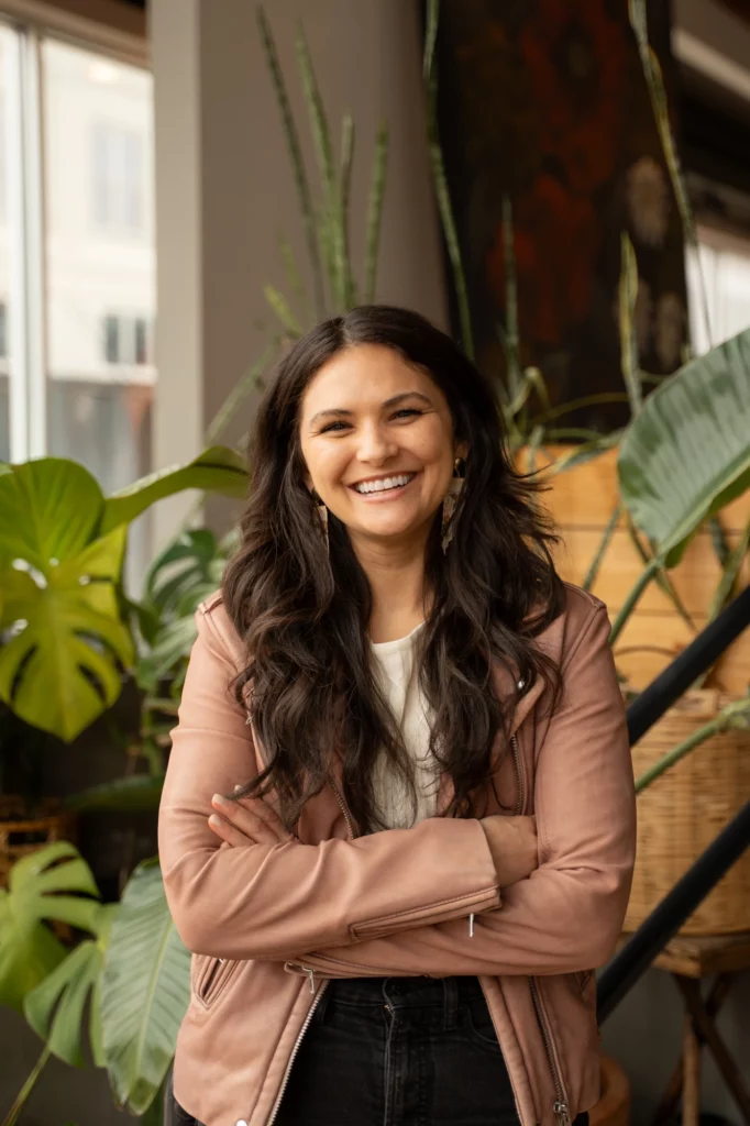 Jessica Crum, CEO, wearing a light pink jacket and white shirt, stands indoors with arms crossed, smiling in front of green plants.