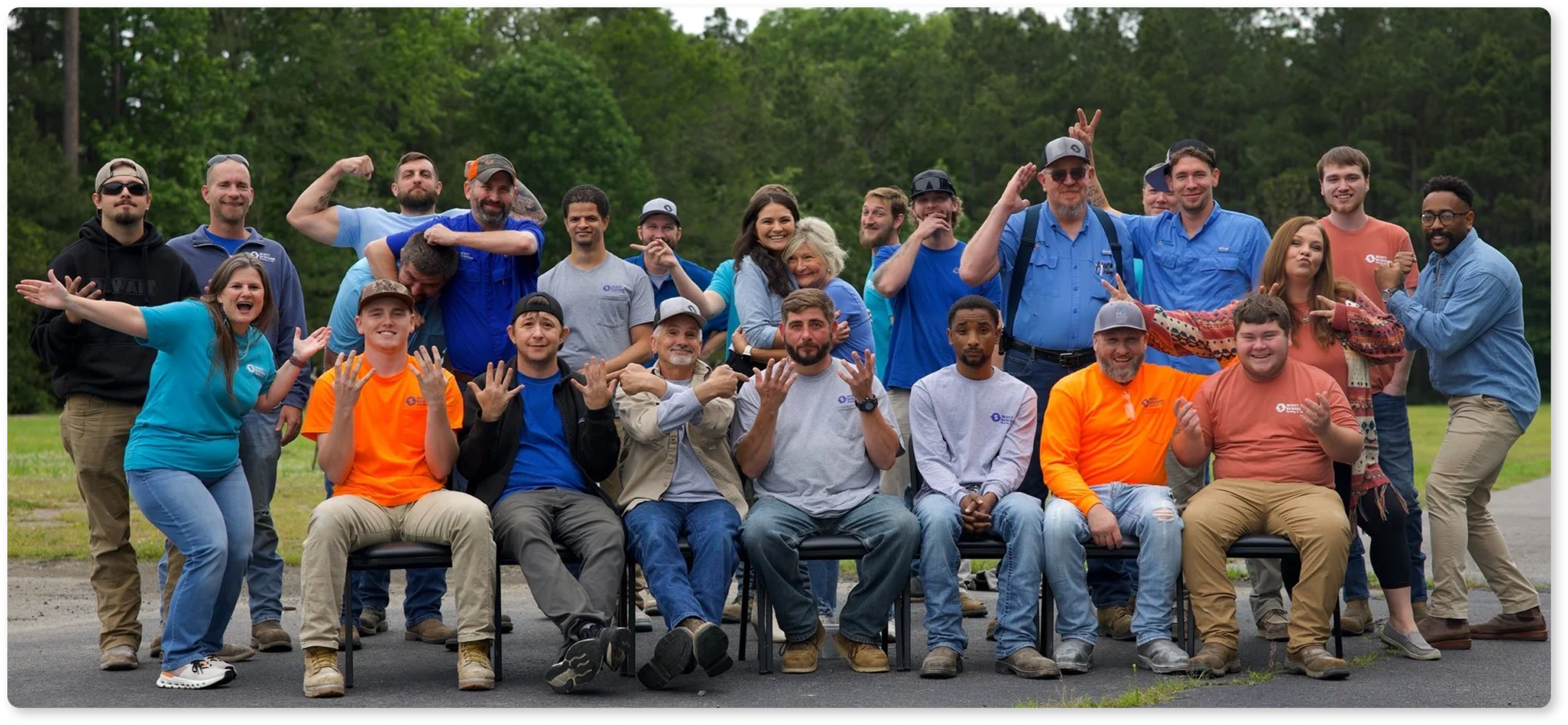 A group of Scott Systems employees, some seated and some standing, pose outdoors, making playful gestures and smiling at the camera.